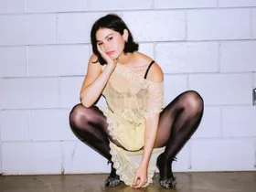 gracie abrams squat poses against tiled wall wearing sheer dress, tights and heels