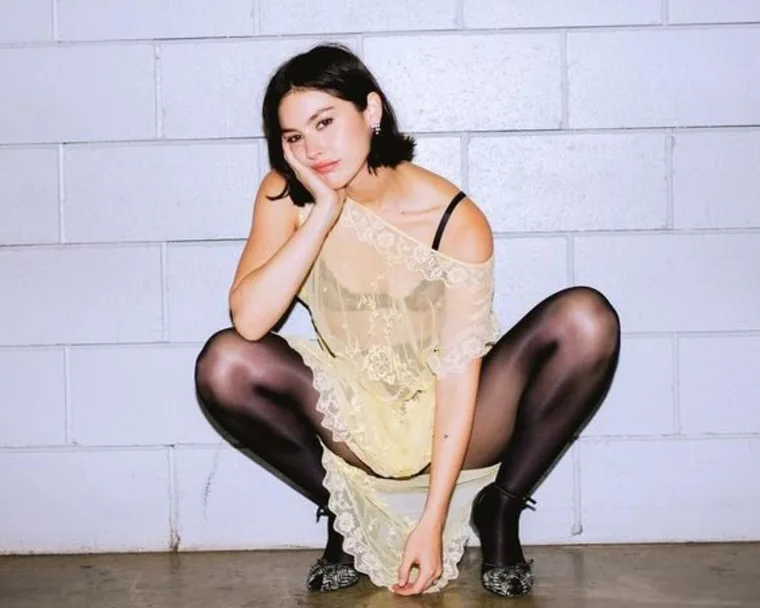 gracie abrams squat poses against tiled wall wearing sheer dress, tights and heels