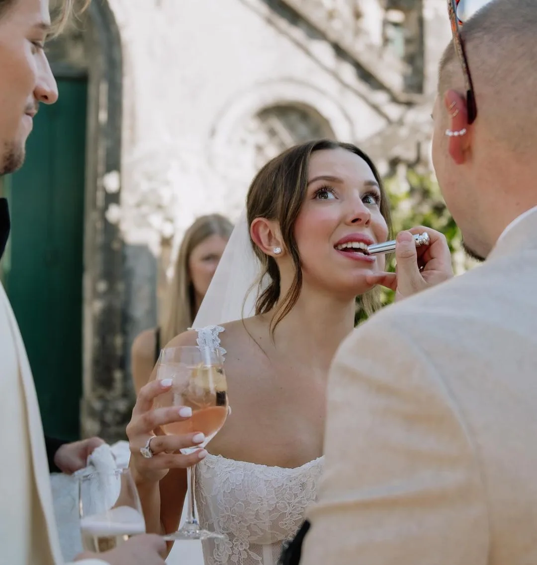 millie bobby brown receiving a wedding makeup touchup with lipstick being applied by her makeup artist