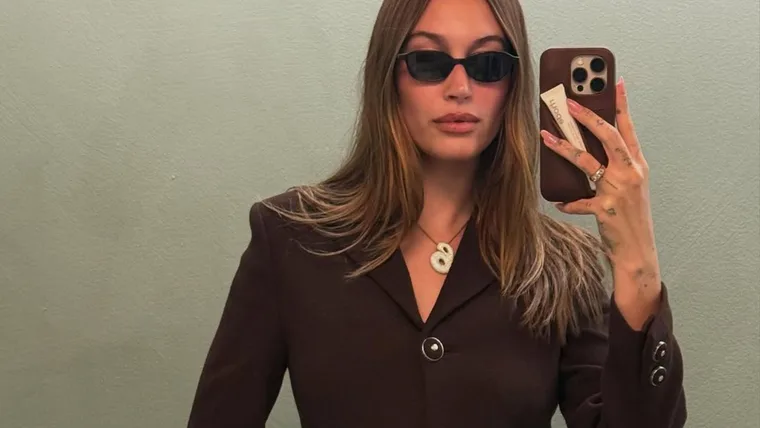 Hailey Bieber taking a selfie wearing her signature Saint Laurent oval shades and a brown suit