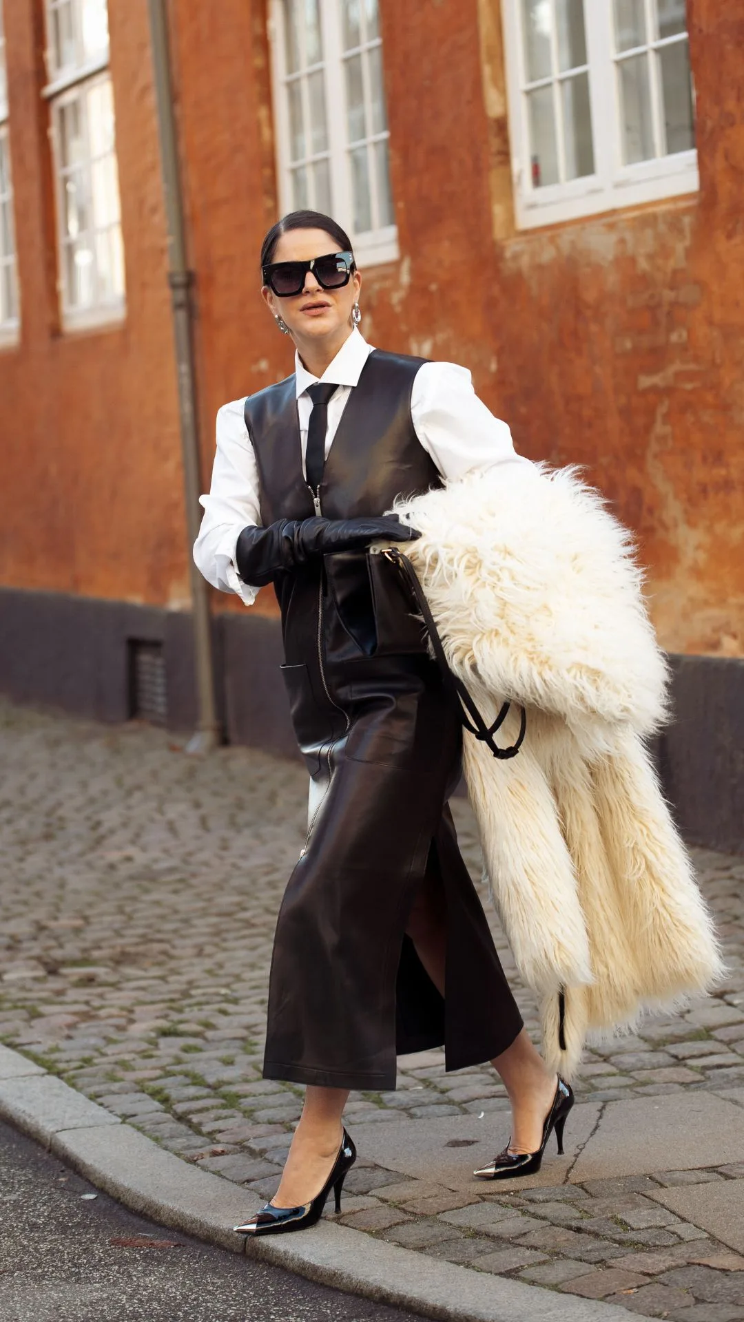 copenhagen street style fashion week autumn winter 2025 woman wears leather skirt, gloves and vest while carrying white faux fur coat