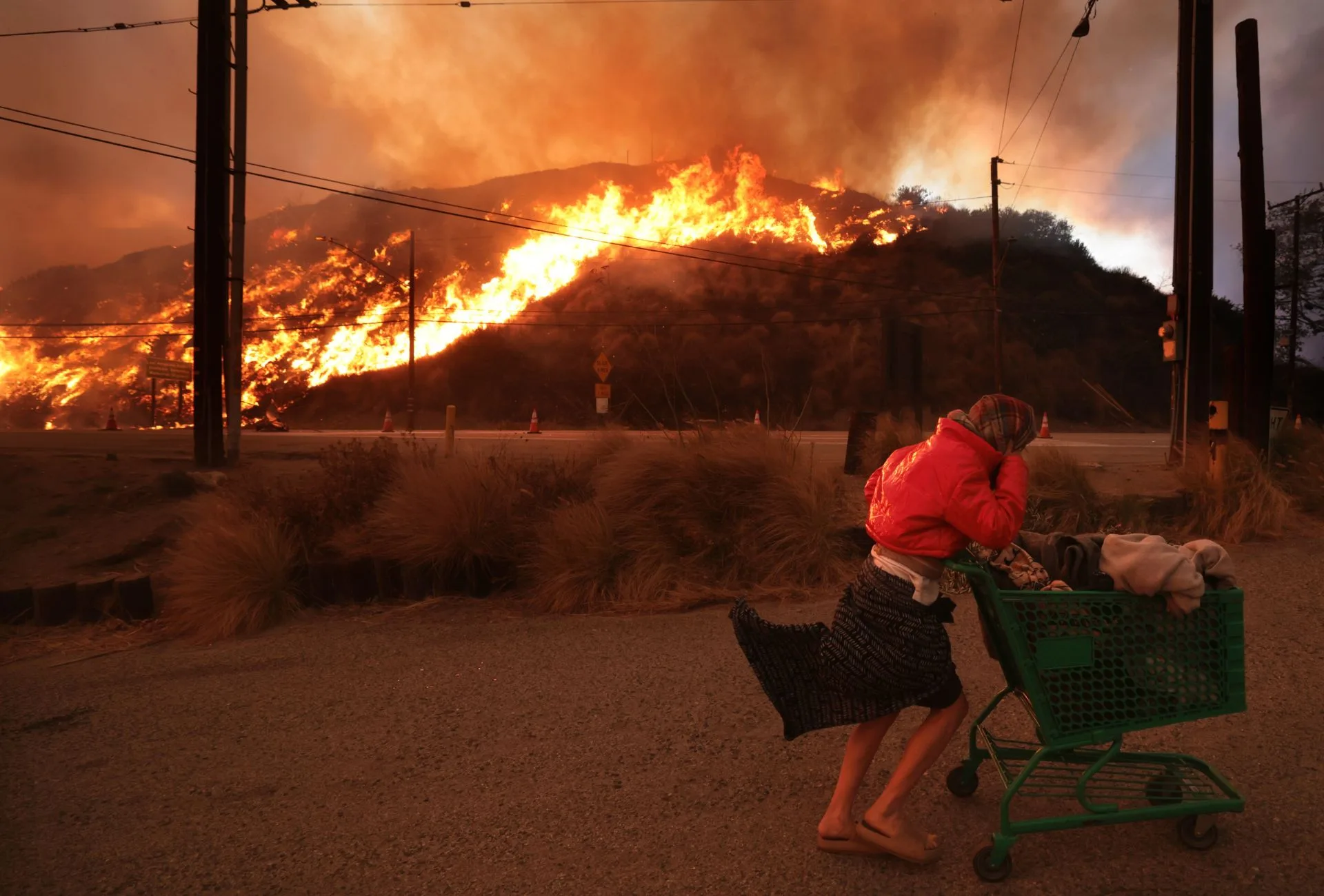 A woman pulling a trolley past a hill that is ablaze during the LA fires