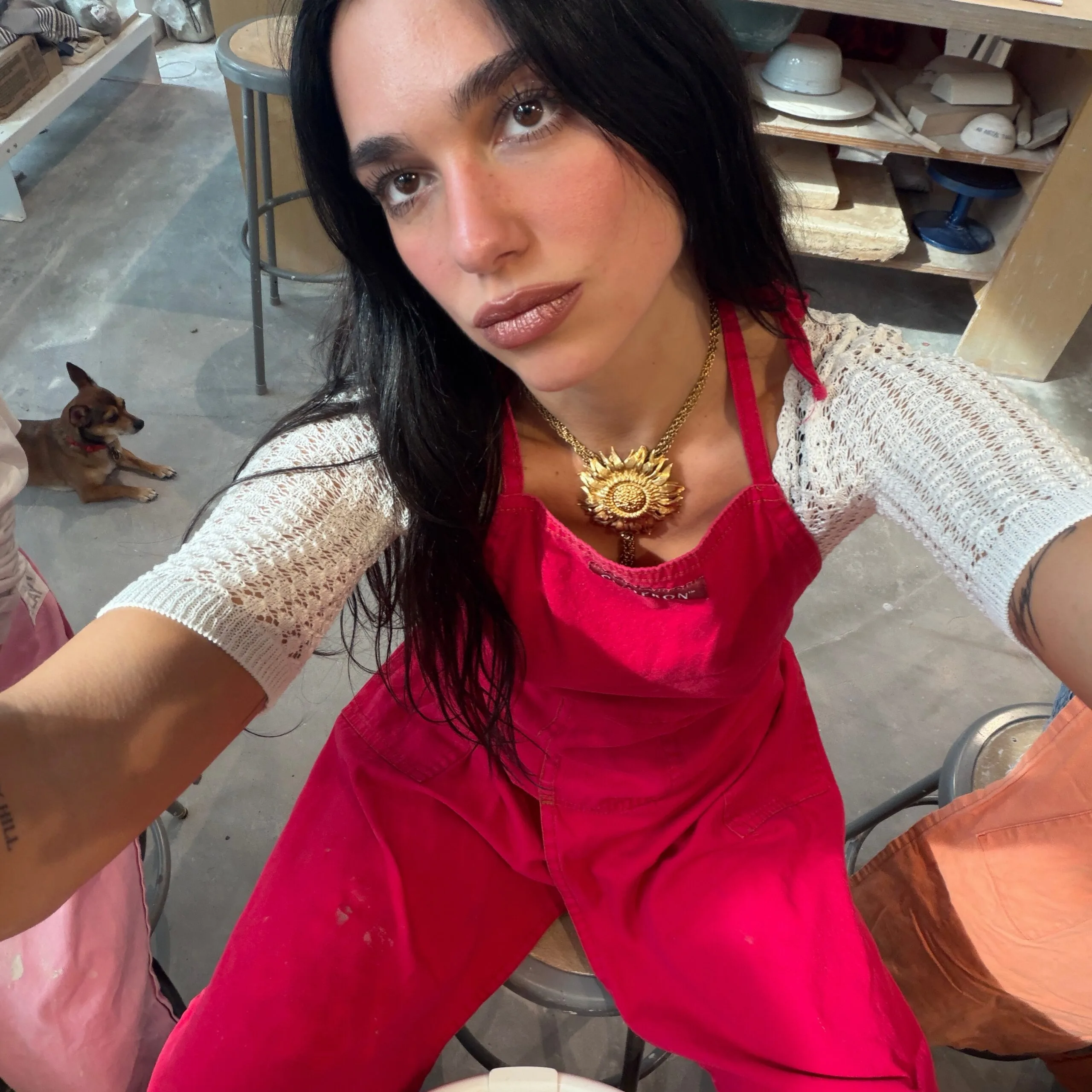Dua Lipa snaps a selfie in pottery class wearing a red apron, gold statement sunflower necklace, and a white With Jean crochet knit top. 