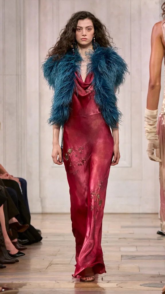 A look from the Ralph Lauren show with the model wearing a crimson slip dress and a teal feather vest.