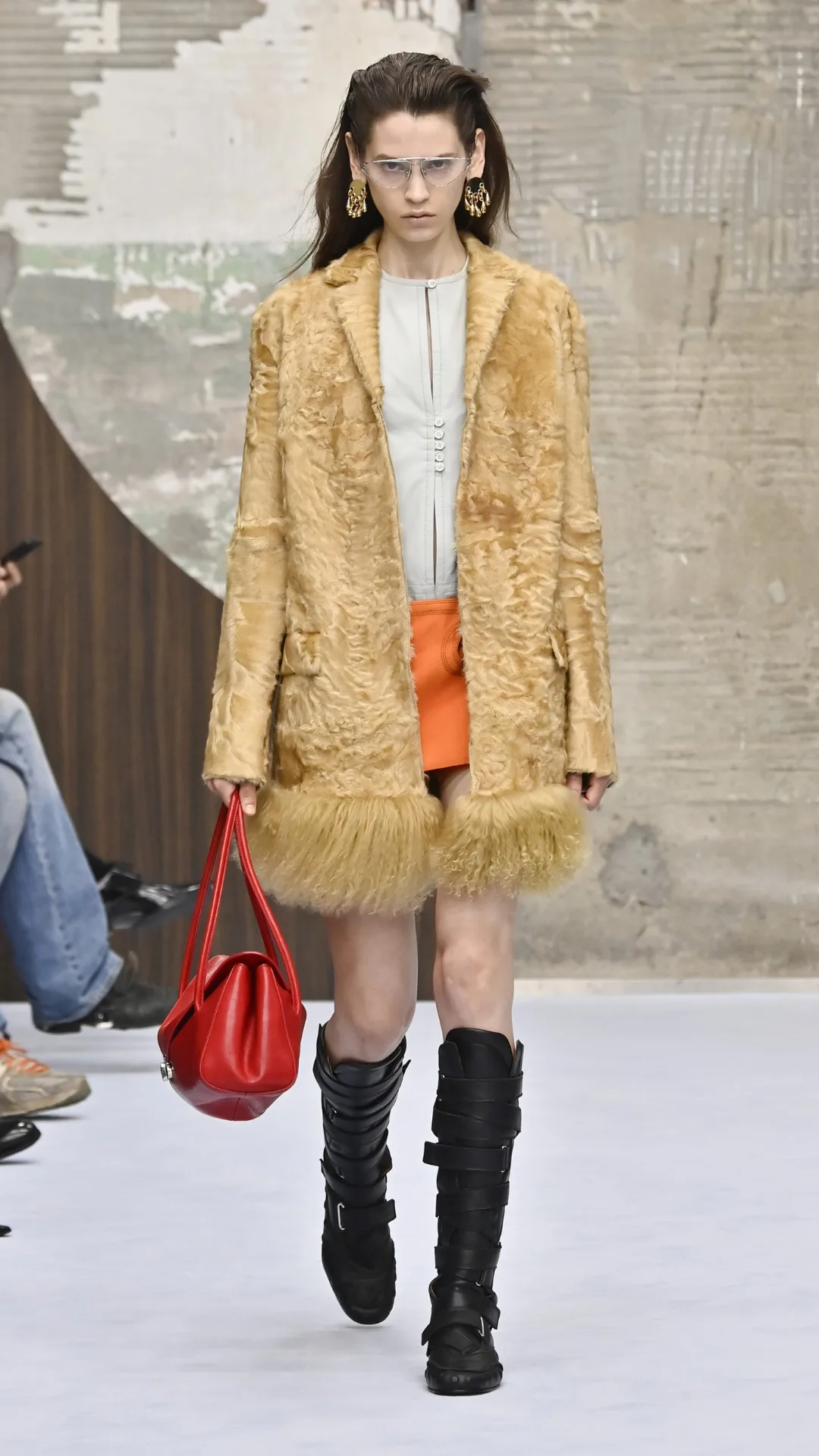 A model in an orange skirt, light blue top and tan coat