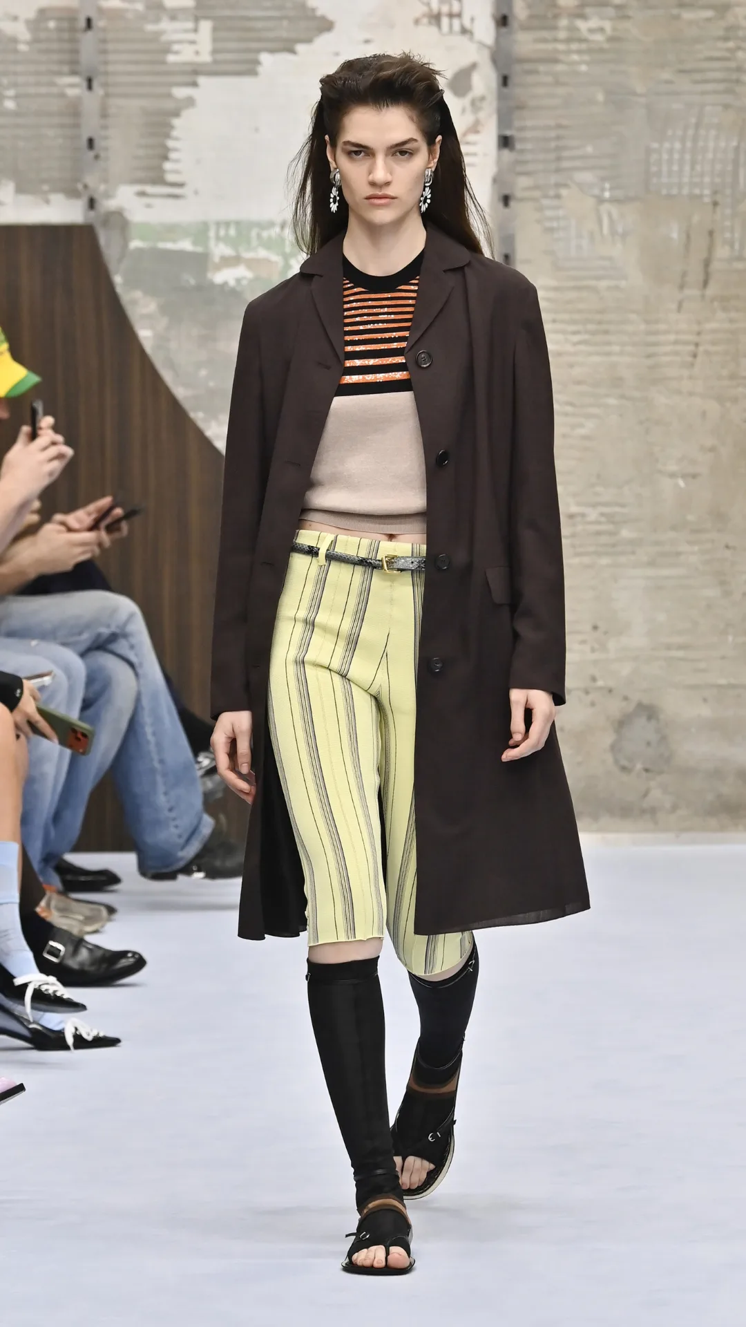A model in yellow capri pants and a brown coat with sweater underneath