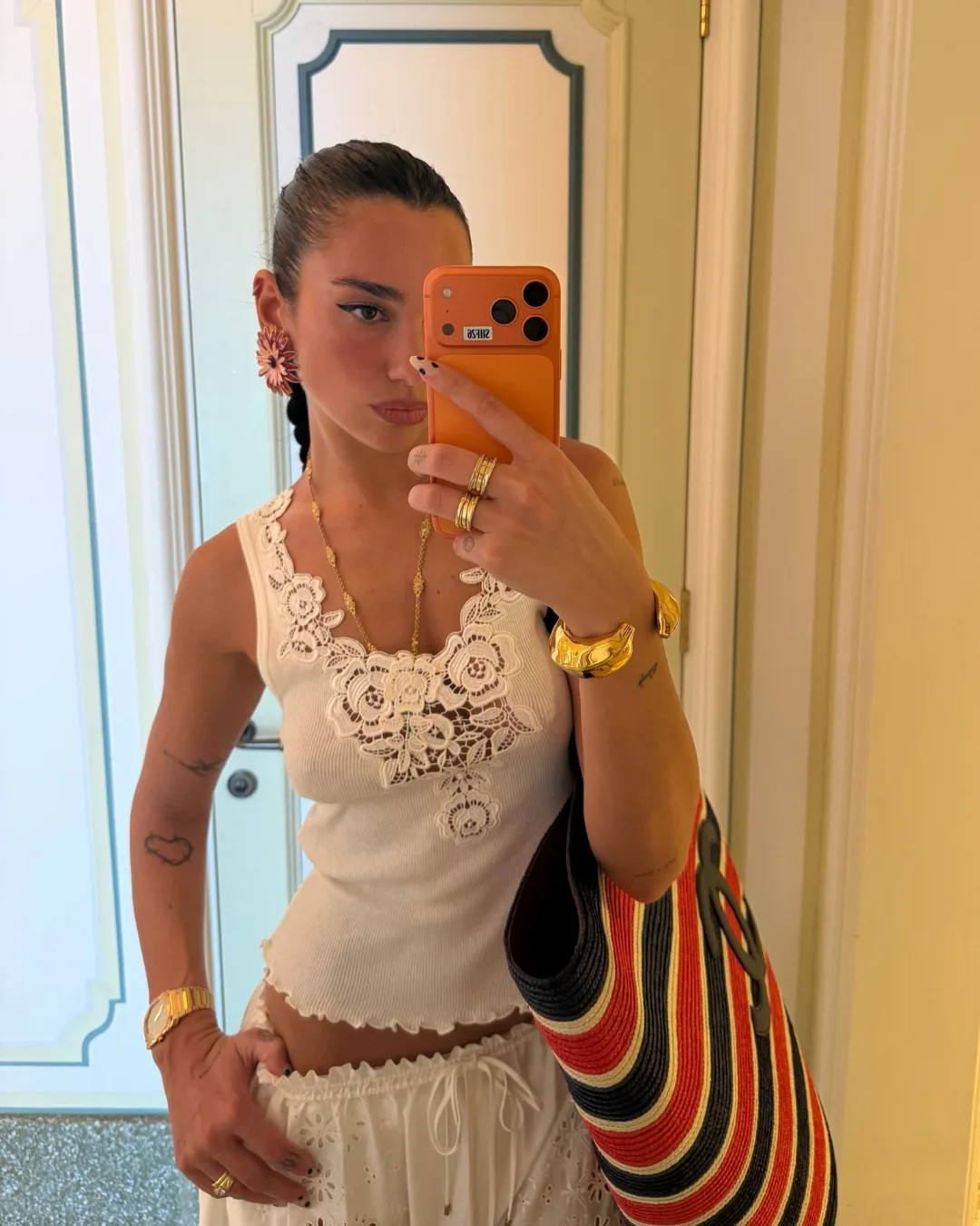 Dua Lipa takes a mirror selfie in a white beach outfit carrying a large blue and red Chanel raffia beach bag