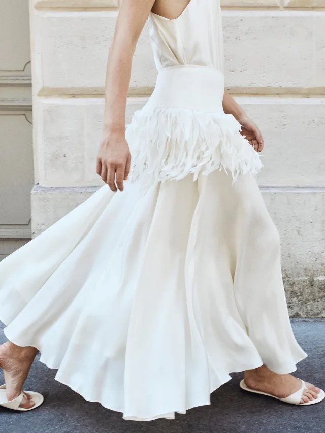Close up of woman wearing long, flowing white maxi skirt belted with a layered feather belt and open-toe white mules from St Agni bridal collection 2027