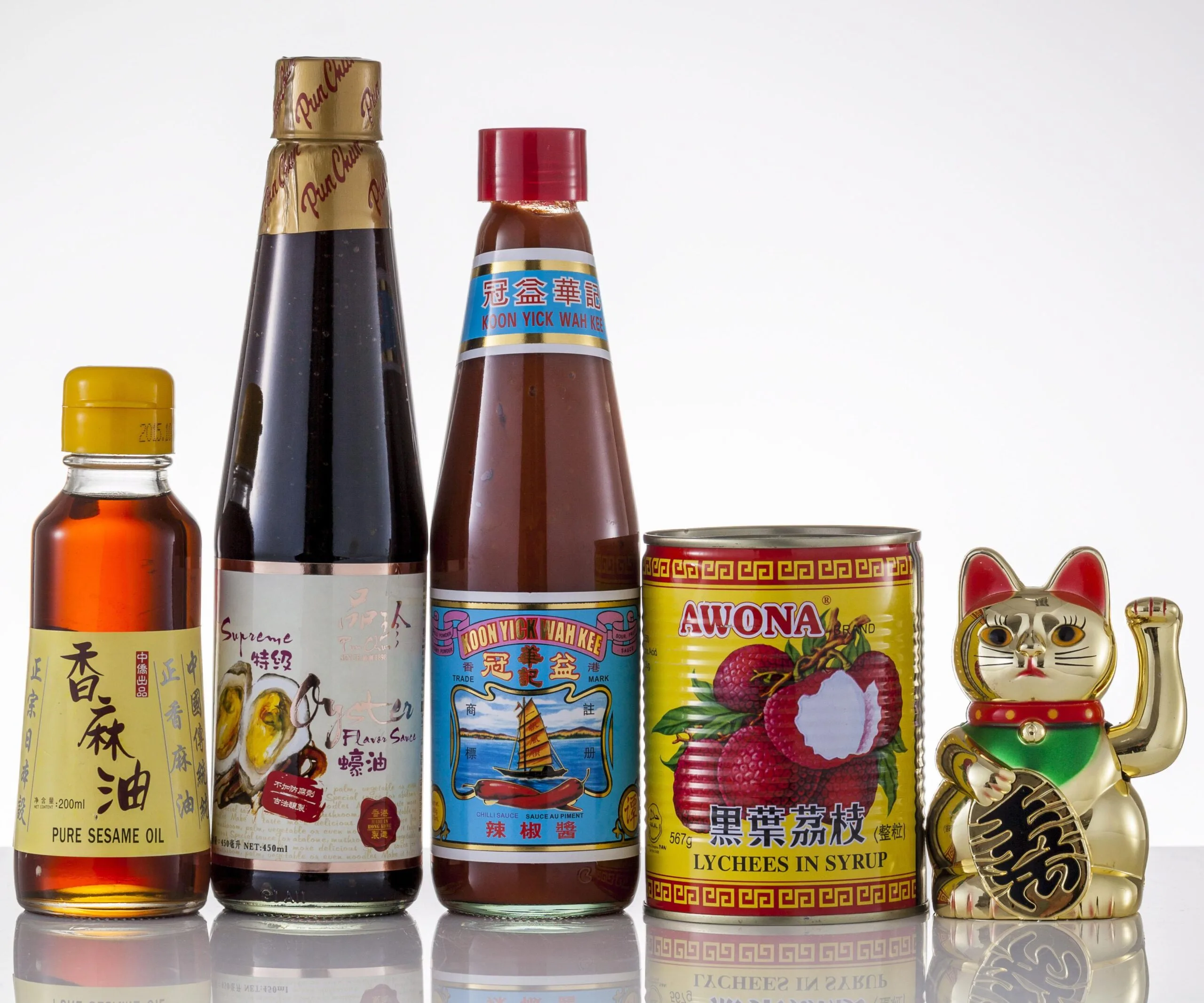 Bottles of sesame oil, oyster sauce, chili sauce, canned lychees, and a gold waving cat figurine on a white background.