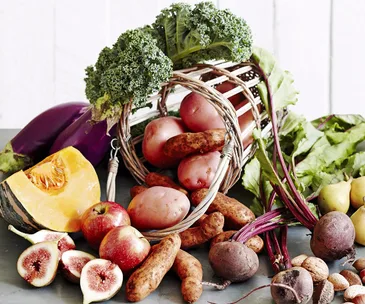A wicker basket spilling over with fresh produce, including potatoes, kale, beets, figs, apples, and a squash slice.