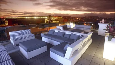 Rooftop lounge with modern white wicker furniture, overlooking city lights at sunset.