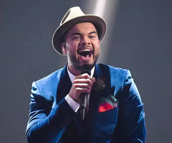 Singer in a blue suit and hat passionately performs on stage with a microphone.