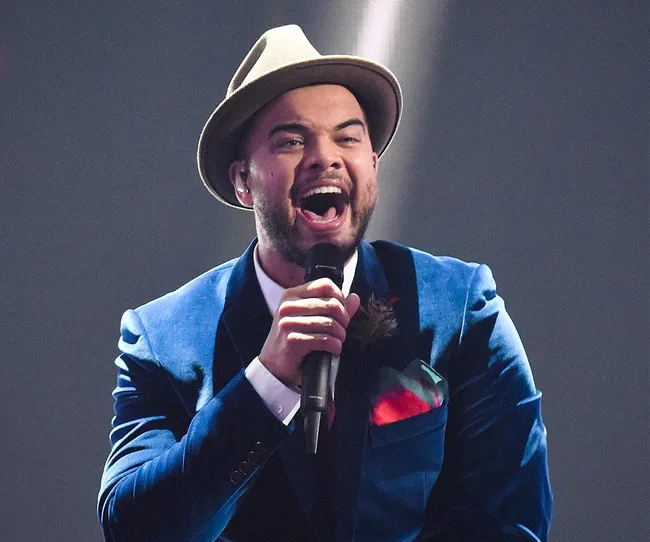 Singer in a blue suit and hat passionately performs on stage with a microphone.
