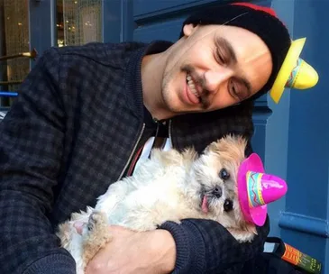 Man cuddles a small dog wearing a pink hat; both look content, with the man also wearing a yellow hat.