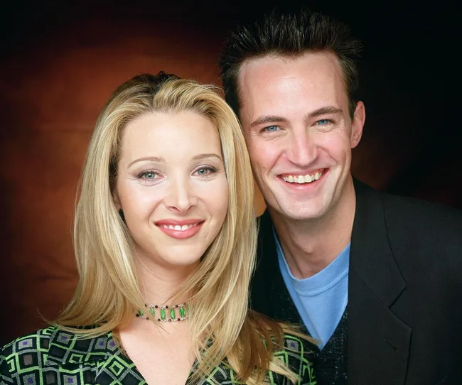 A man and a woman smiling together, posed against a blurred brown background.