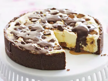 Chocolate and caramel cheesecake with a slice missing on a white cake stand.