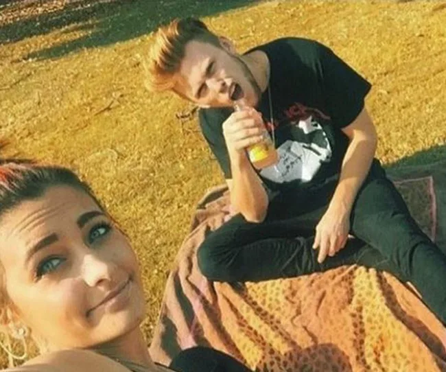 Two people sitting on a blanket outdoors; one holding a drink, both enjoying a sunny day.
