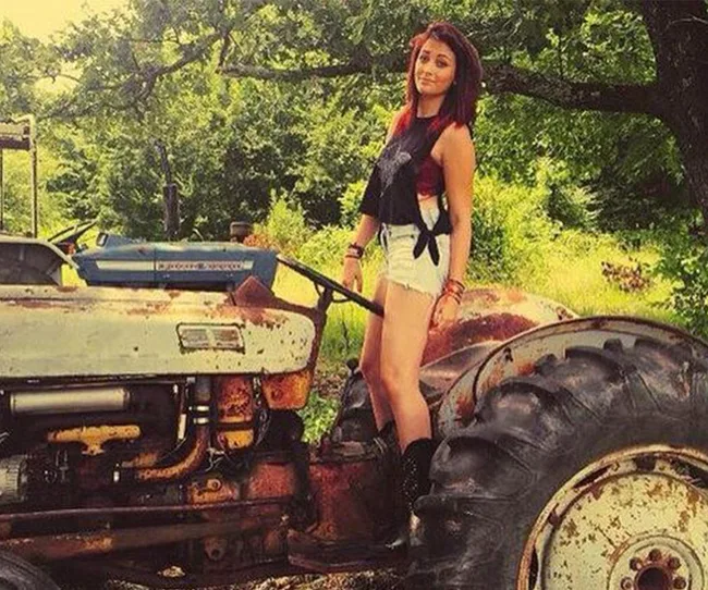 Person standing on a rustic tractor in a lush, green outdoor setting, wearing a black top and denim shorts.
