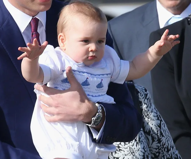 A baby in a white outfit being held by an adult in a blue suit, raising its arms forward.