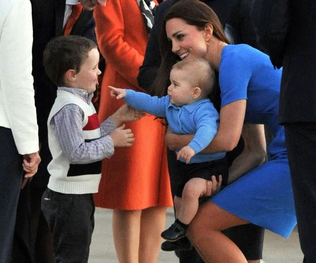 A woman in blue crouches, holding a baby who points at a smiling young boy in a vest and shirt.