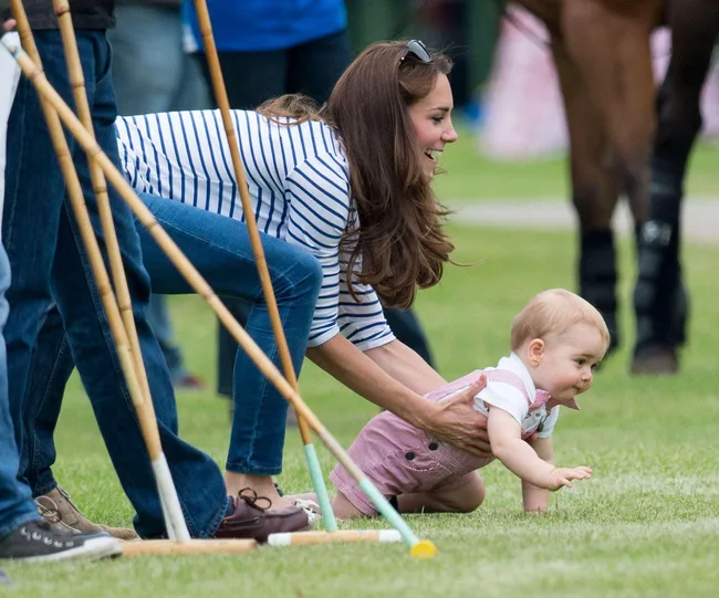A woman in a striped shirt helps a crawling baby on grass near polo mallets.