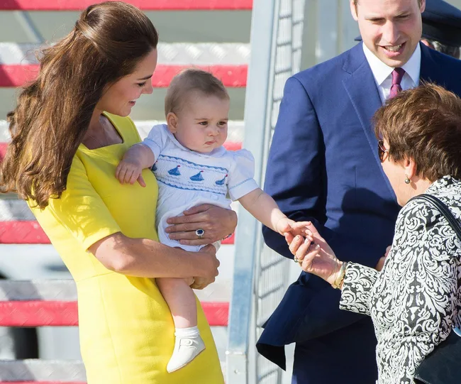 A woman in a yellow dress holding a baby while another adult interacts with them on a sunny day.