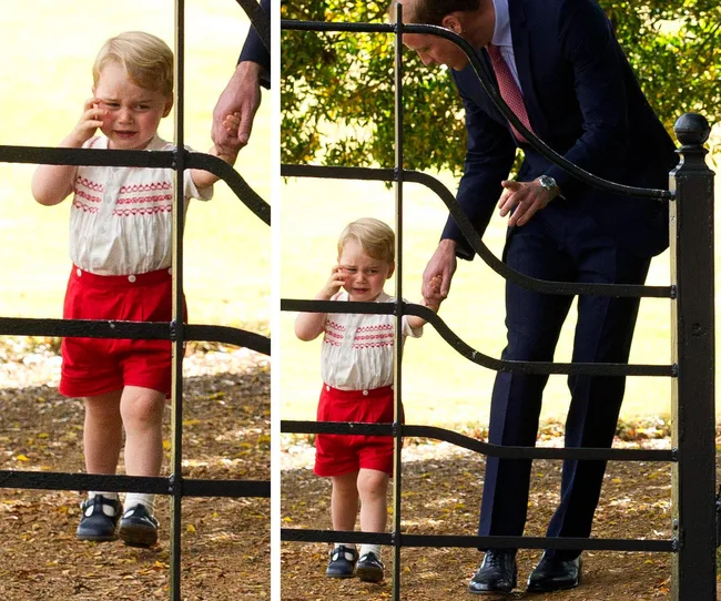 A young child in red shorts, crying while holding the hand of a man in a suit by a metal gate outdoors.