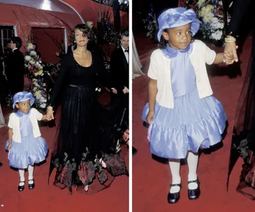 A young girl in a blue dress holds hands with an adult on a red carpet, smiling and wearing a matching hat.