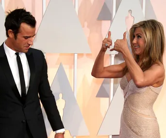 Two people at a formal event, one in a tuxedo looking down and the other giving a thumbs up sign while smiling.