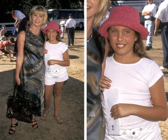 A woman in a patterned dress stands with a young girl in a pink hat and white outfit outdoors, both smiling at the camera.