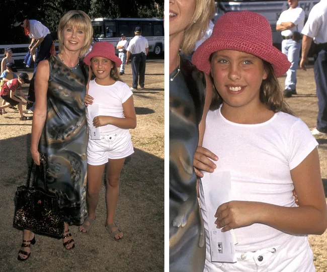 A woman in a patterned dress stands with a young girl in a pink hat and white outfit outdoors, both smiling at the camera.