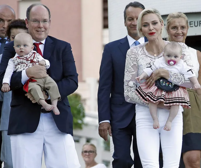 A man and woman smiling, each holding a baby, with two other adults in the background outdoors.