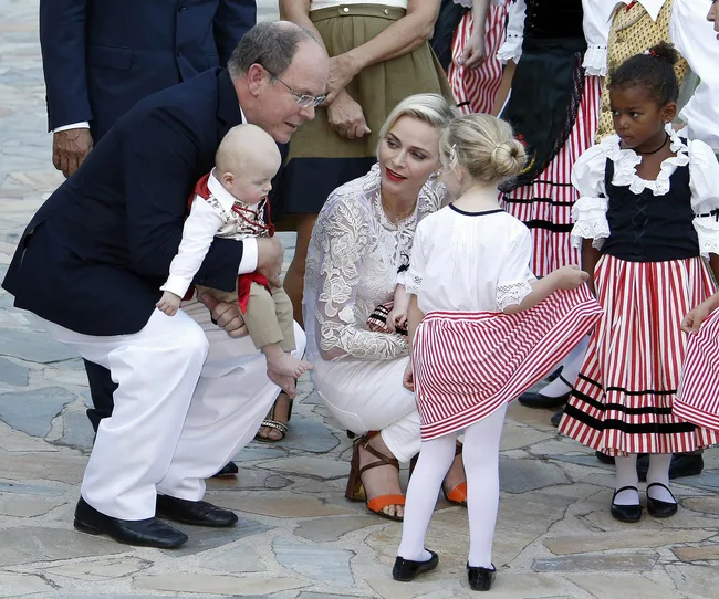 A man holding a baby crouches next to a woman and children in traditional striped costumes, outdoors on a stone path.