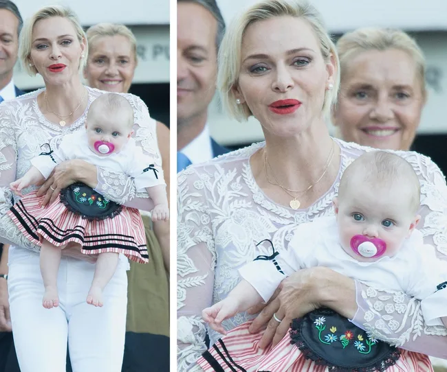 A woman in a white lace top holds a baby with a pink pacifier and striped dress, surrounded by smiling people.