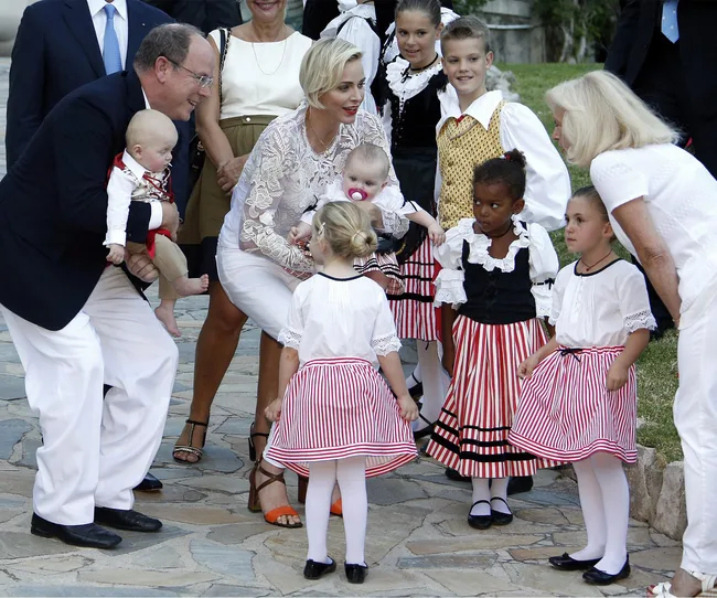 A group of adults and children, some in traditional striped dresses, gather together outdoors on a stone path.