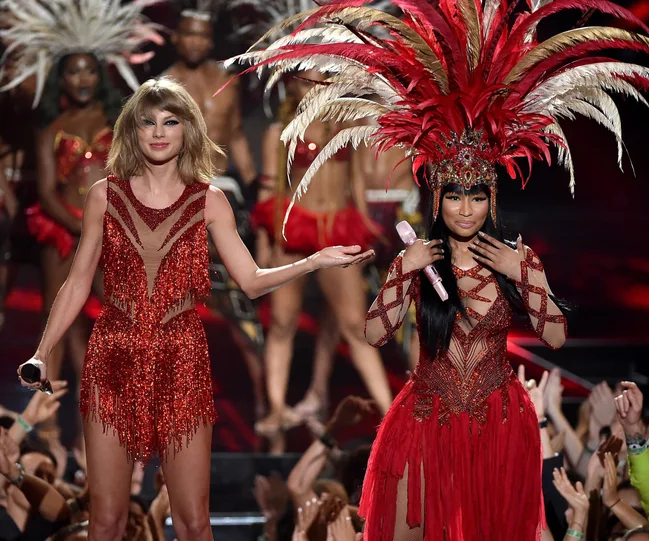 Two female performers in red costumes on stage, surrounded by dancers with feathered headdresses, during an award show.