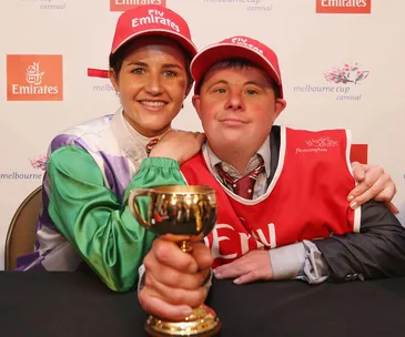 A jockey and companion smiling, holding a Melbourne Cup trophy, both wearing red Emirates hats at a press event.