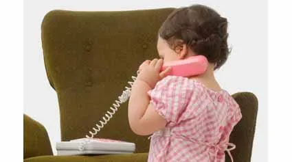 A toddler in a pink checkered dress talking into a pink toy phone while standing beside a green armchair.