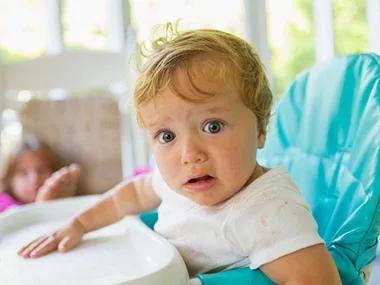 Baby with curly hair in a high chair, looking surprised, wearing a white shirt; a blurry person in the background.