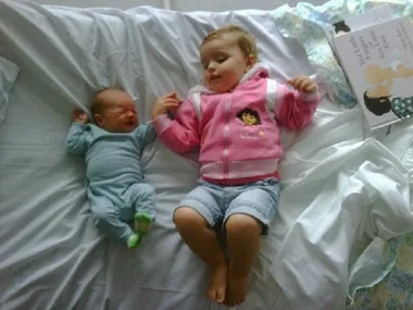A toddler and crying baby lie on bed holding hands, with a parenting book nearby.