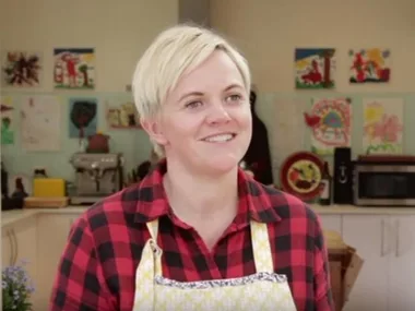 A person in a kitchen wearing a red plaid shirt and apron, smiling. Kitchen utensils and artwork are visible in the background.