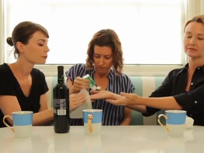 Three women sitting at a table with mugs; one cleans another's hand with a wipe, resembling playful parenting interaction.