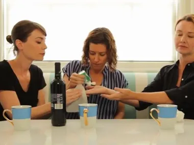 Three women sitting at a table with mugs; one cleans another's hand with a wipe, resembling playful parenting interaction.