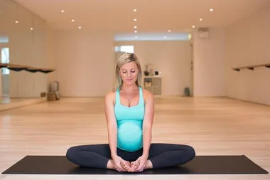 A pregnant woman practicing a seated yoga pose in a bright studio, wearing a teal top and black leggings.