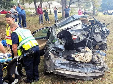 "Rear of heavily damaged car in crash; emergency responders at scene, crowd of onlookers in background."