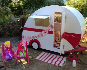 Mini white and red caravan with "Maison Amour" banner, two colorful chairs, set in a garden patio.