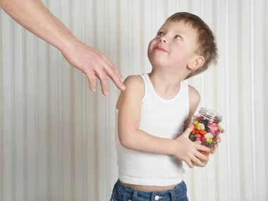 A child holding a jar of colorful candies looks up, as an adult hand reaches out towards them.