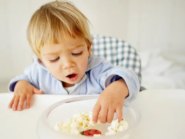 Toddler reaching for popcorn in a bowl, highlighting potential choking hazards for young children.