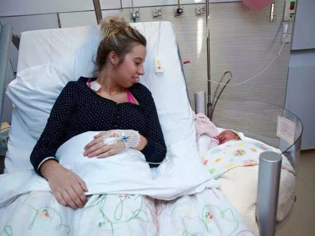 Mother in hospital bed smiles at newborn in bedside crib.