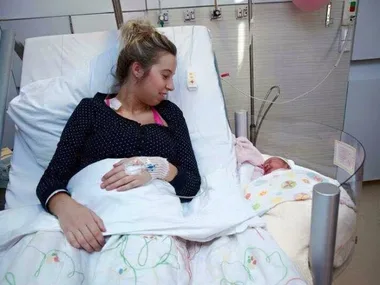 Mother in hospital bed smiles at newborn in bedside crib.